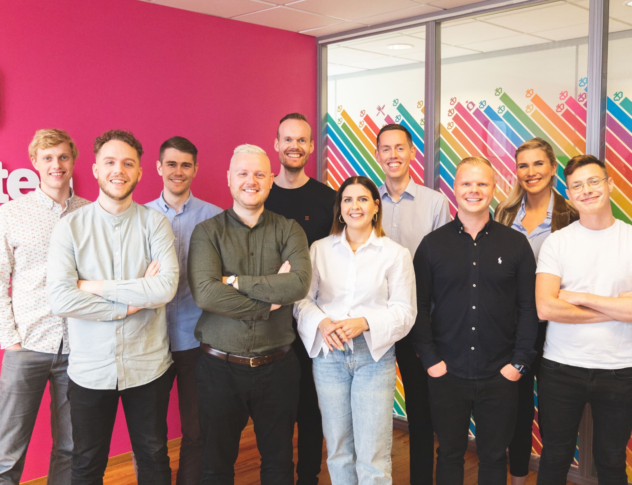 The Smitten team smiling in front of the office’s pink wall and rainbow-striped glass